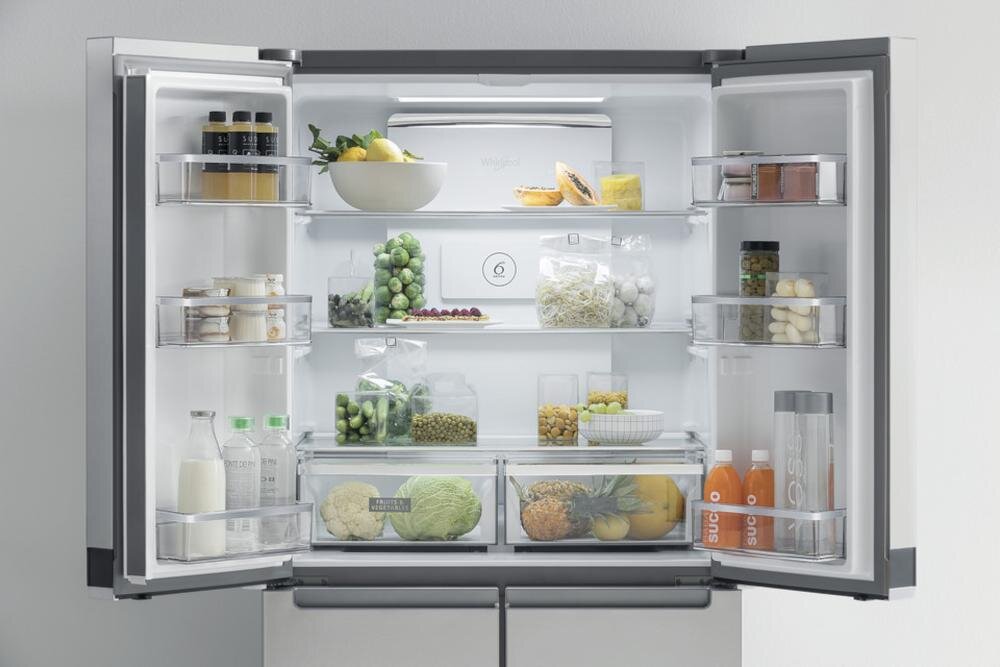 Otwarta lodówka z widocznymi półkami wypełnionymi różnorodnymi produktami spożywczymi, takimi jak owoce, warzywa, napoje i słoiki. Na tylnej ścianie widoczne jest logo \'whirlpool\'.