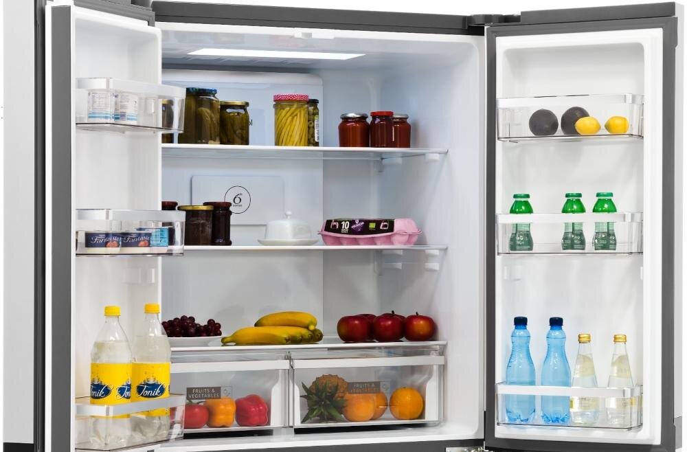 Otwarta lodówka z widocznymi półkami wypełnionymi różnymi produktami spożywczymi, takimi jak słoiki, butelki, owoce i napoje. Wnętrze jest dobrze oświetlone, a na drzwiach znajdują się dodatkowe półki z napojami i cytrusami.