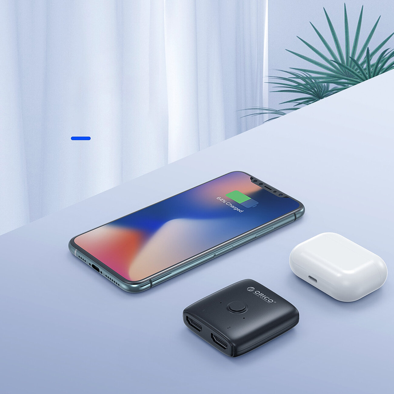 Na jasnym tle znajduje się smartfon z ikoną ładowania, czarne urządzenie z logo \'Orico\' oraz białe etui. W tle widoczna jest roślina i zasłona.