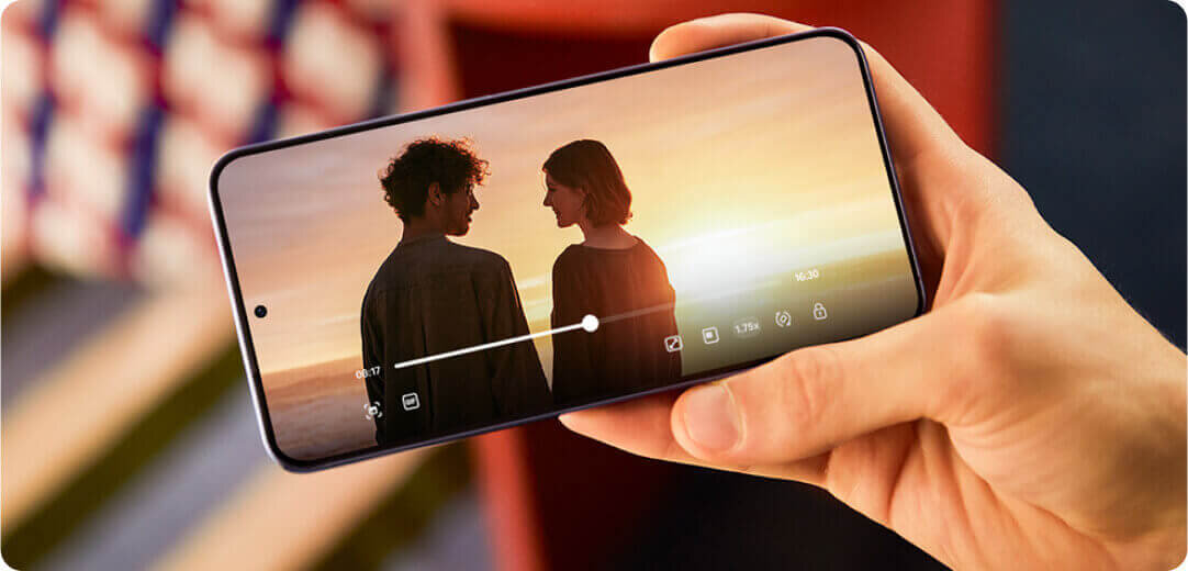 Osoba trzyma smartfon, na ekranie którego widać zdjęcie dwóch osób na tle zachodzącego słońca. Na ekranie widoczne są również ikony interfejsu użytkownika, w tym pasek przewijania i symbole aplikacji.