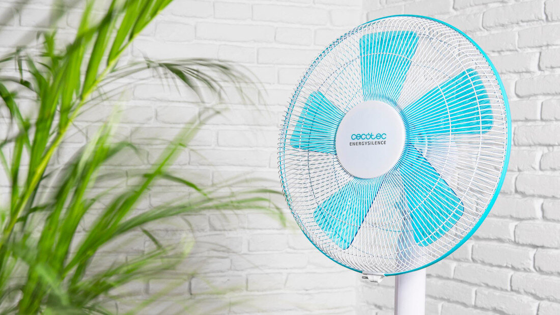 Wentylator z niebieskimi łopatkami i białą siatką ochronną, na tle białej ceglanej ściany. Na środku wentylatora widoczny jest napis \'cecotec EnergySilence\'.