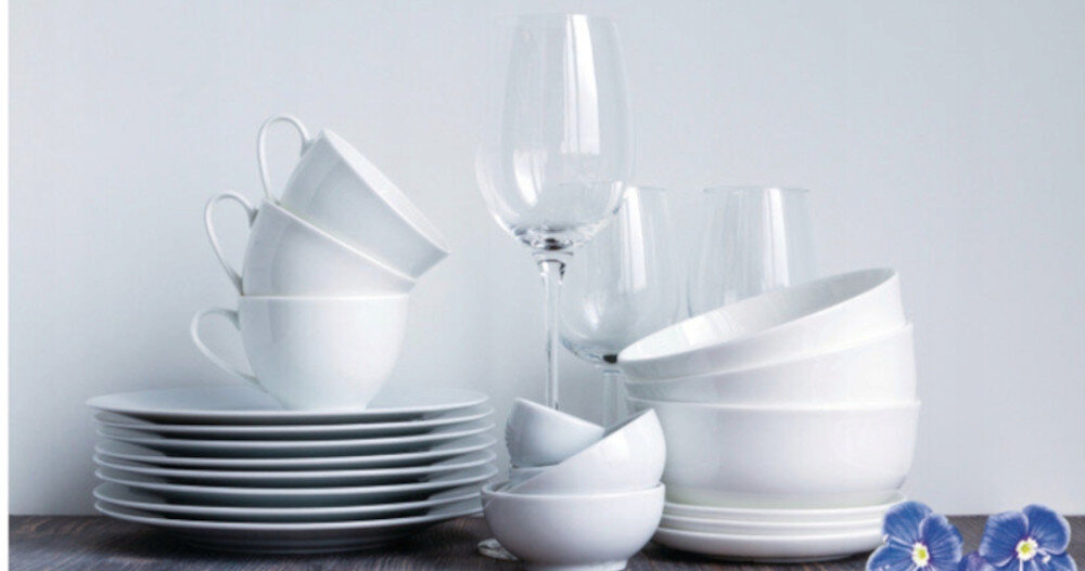 Na obrazie widoczny jest zestaw białej porcelany, w tym talerze, filiżanki i miski, ustawione na drewnianej powierzchni. Obok znajdują się kieliszki do wina oraz niebieskie kwiaty.