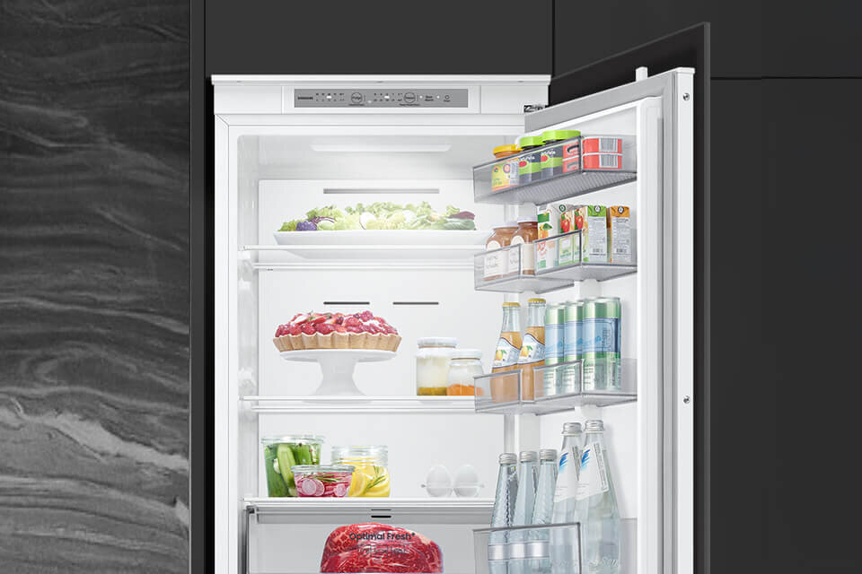 Otwarta lodówka z widocznymi półkami wypełnionymi różnorodnymi produktami spożywczymi, takimi jak warzywa, napoje i ciasto. Na drzwiach lodówki znajdują się dodatkowe półki z kartonami i butelkami.