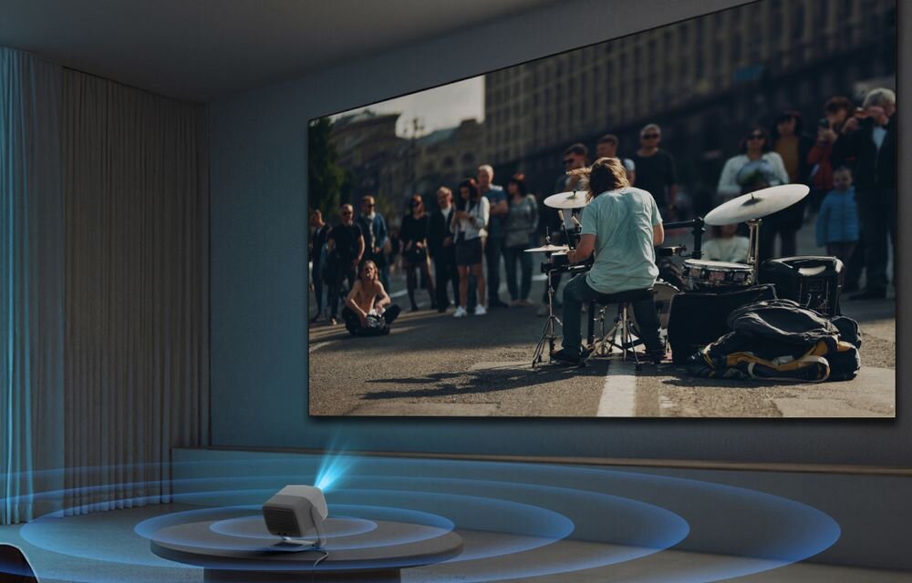 Projektor laserowy JMGO N3 SE JMGO Master Sound dwa głośniki 5 W Dolby Audio 55 Hz, projektor stojący na stoliku wyświetlający obraz na ekranie projekcyjnyym