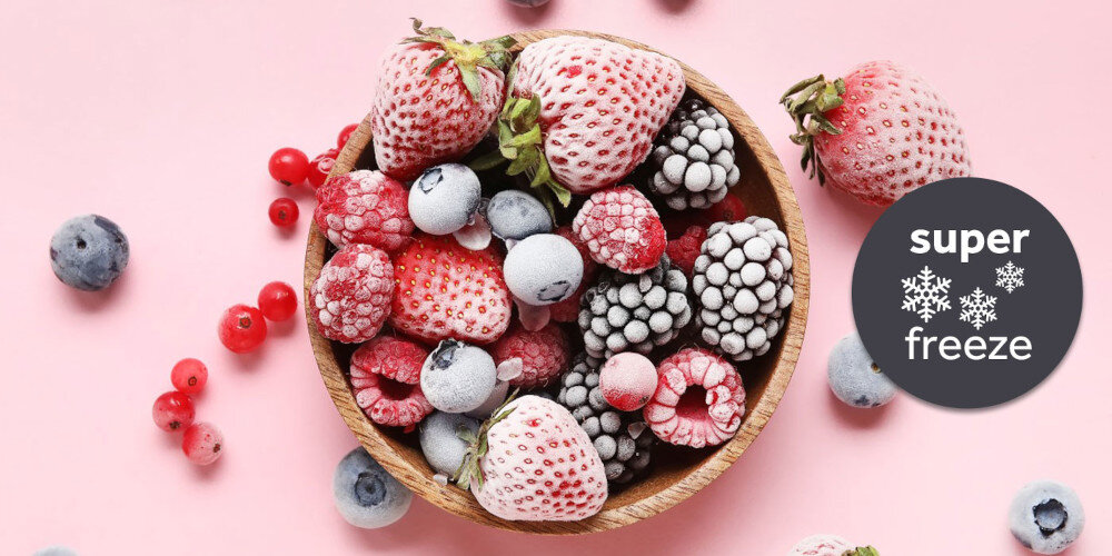 Na różowym tle znajduje się miska z mrożonymi owocami, takimi jak truskawki, maliny i jagody. Obok widoczny jest okrągły znak z napisem \'super freeze\' oraz trzema śnieżynkami.