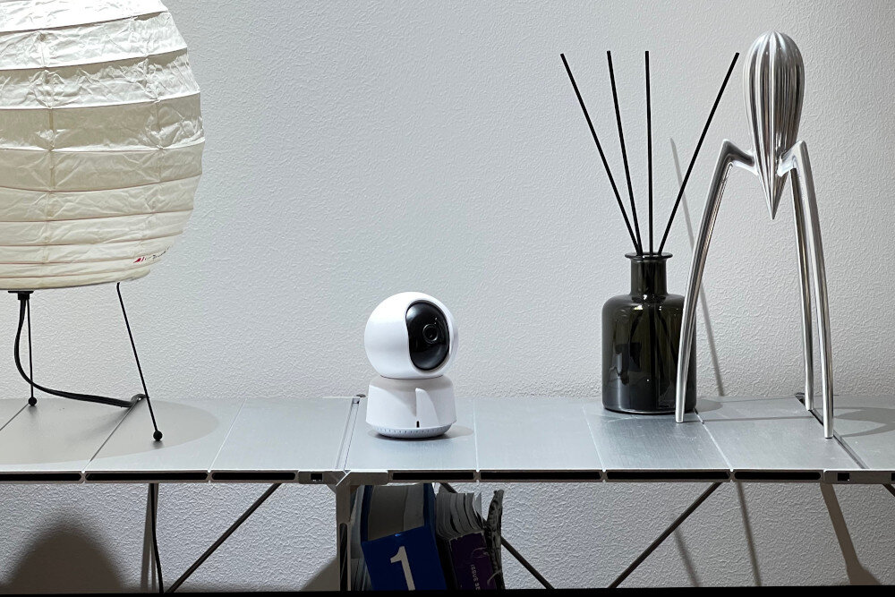 Na stole znajduje się biała, obrotowa kamera oraz czarna butelka z patyczkami. Obok widoczna jest lampa z papierowym kloszem i metalowy przedmiot o futurystycznym kształcie.