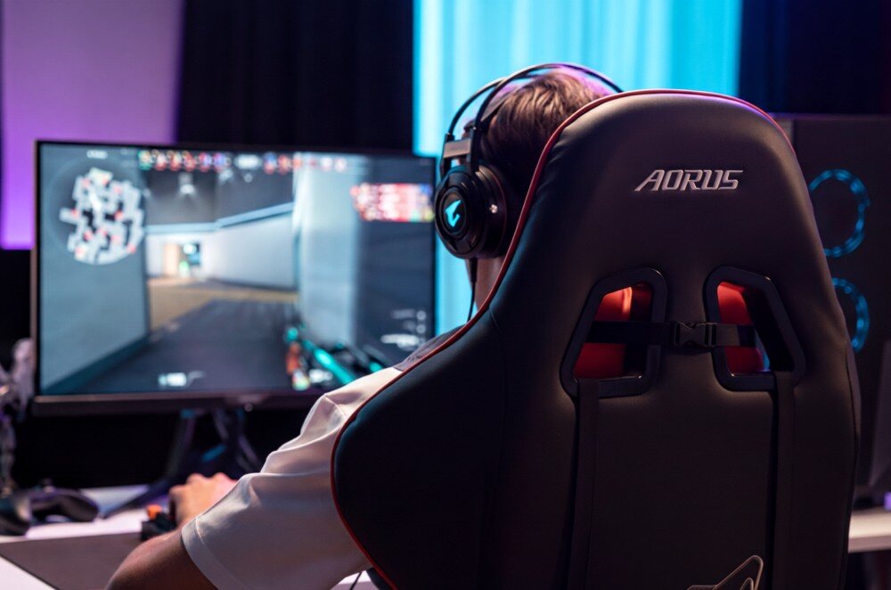 Komputer GIGABYTE Aorus Prime 5 Gracz w słuchawkach siedzący na fotelu z napisem 'AORUS' przed monitorem z widoczną grą