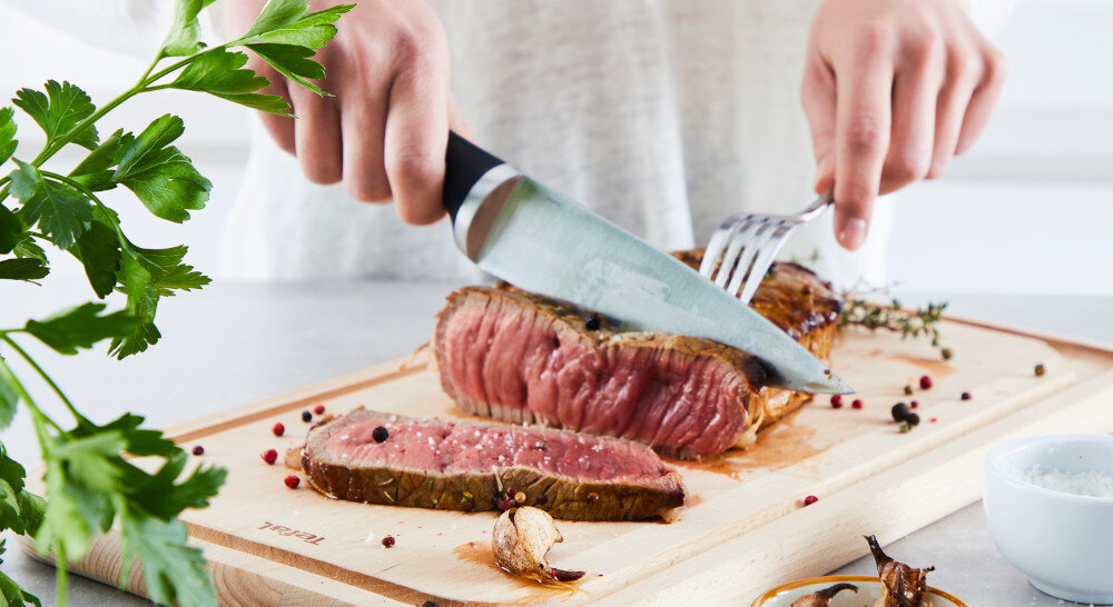 Osoba kroi kawałek mięsa na desce do krojenia, obok leży gałązka ziół i kilka ziaren pieprzu. Na desce widoczne jest logo \'TEFAL\'.