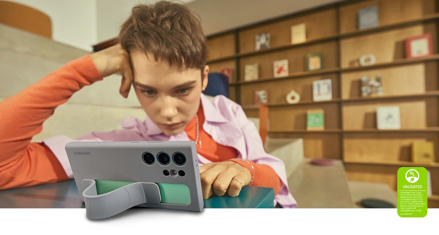 standing grip case z wygodną podstawką Osoba siedzi przy stole, patrząc na telefon w etui z podpórką. W tle widać półki z książkami i ozdobami.