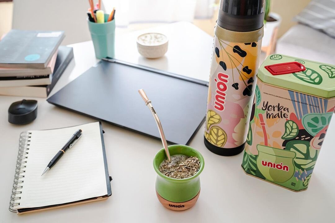 Na biurku znajduje się zestaw do yerba mate z kubkiem, bombillą i puszką z napisem \'Yerba mate union\'. Obok leży otwarty notes z długopisem oraz kilka książek i laptop.