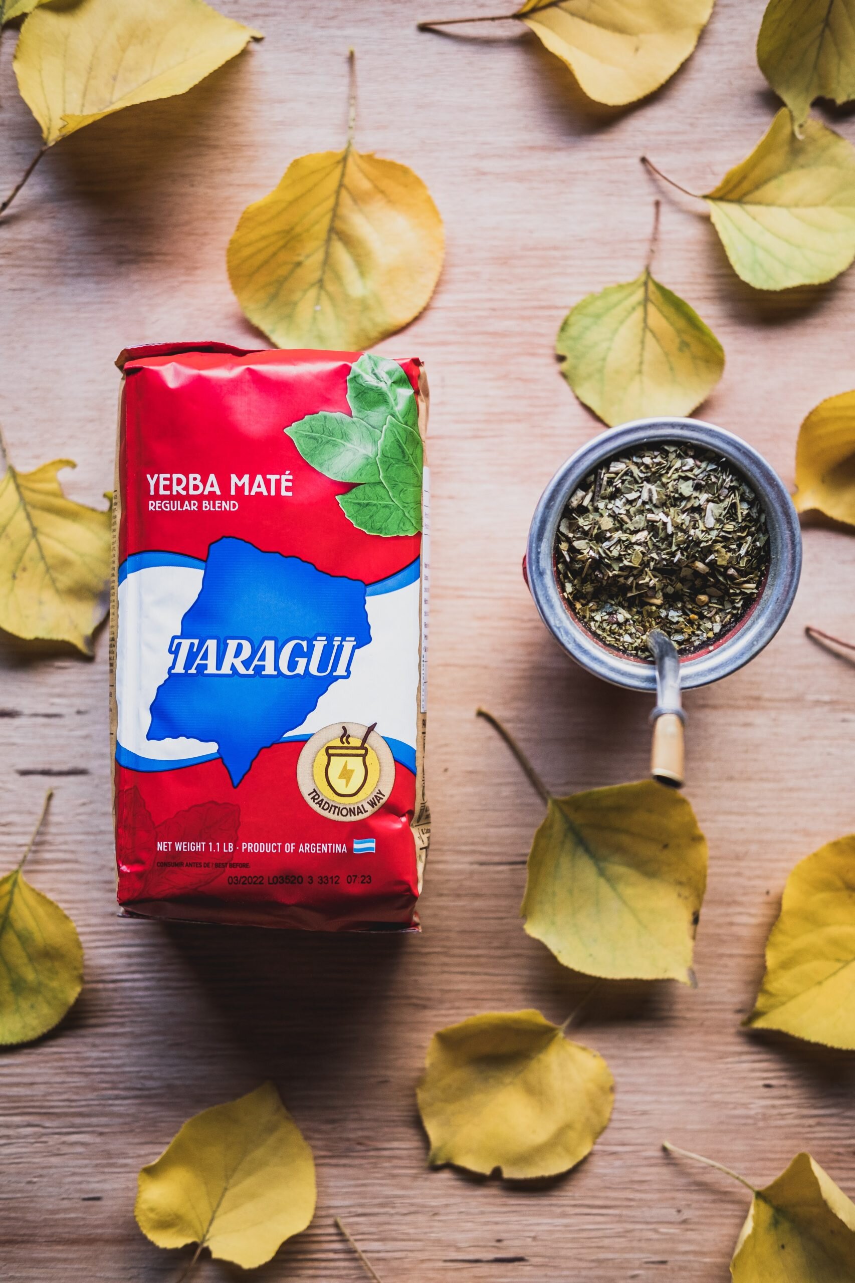 Na drewnianym tle znajduje się opakowanie Yerba Mate Taragui oraz naczynie z suszem. Wokół leżą żółte liście.
