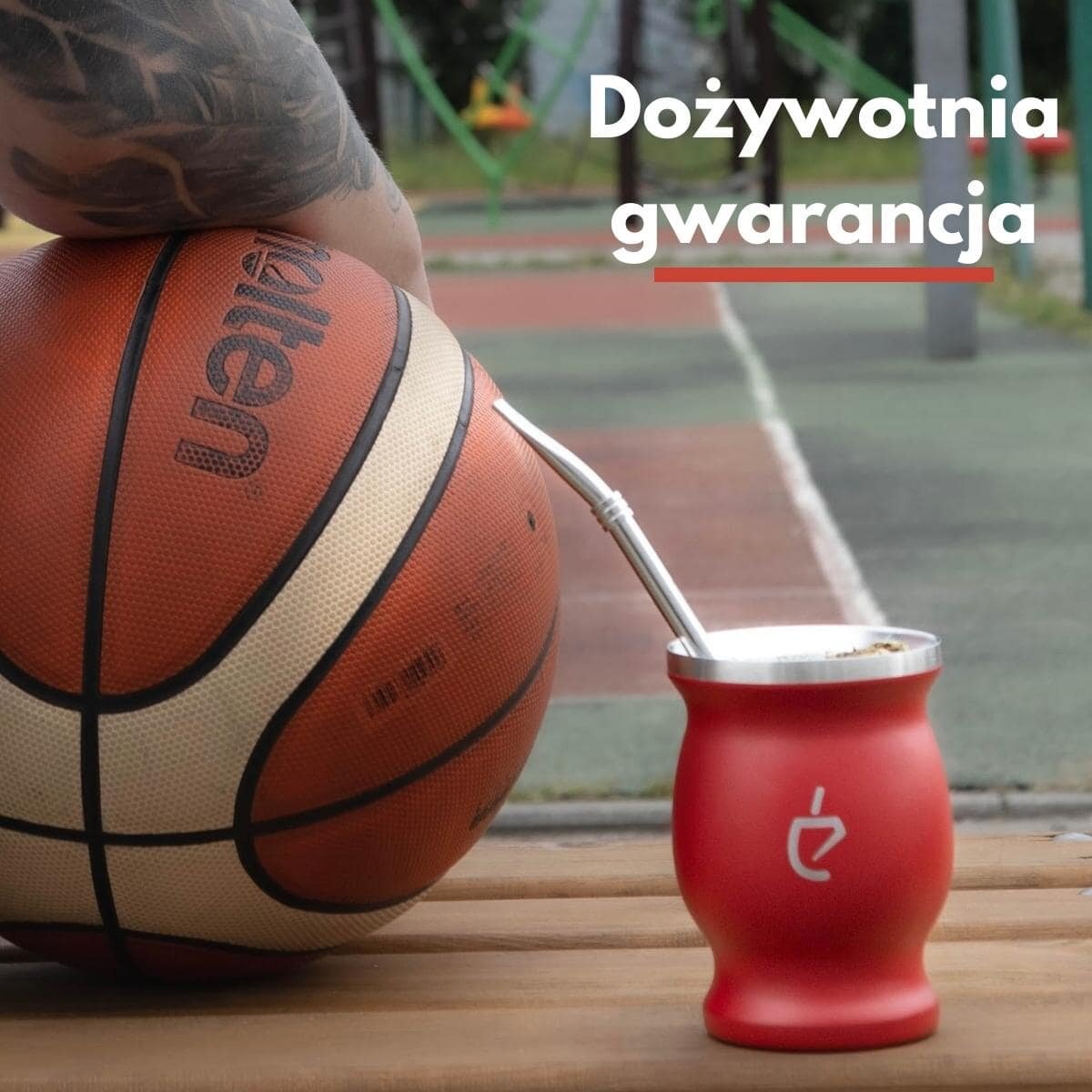 Na drewnianej ławce znajduje się czerwona tykwa z metalową bombillą oraz piłka do koszykówki z widocznym logo. W tle widoczny jest napis \'Dożywotnia gwarancja\'.