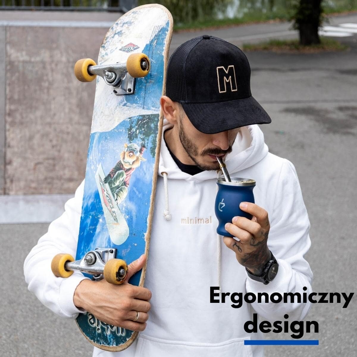 Osoba w białej bluzie i czapce z literą \'M\' trzyma deskorolkę oraz pije z naczynia do yerba mate. Na obrazie widoczny jest napis \'Ergonomiczny design\'.