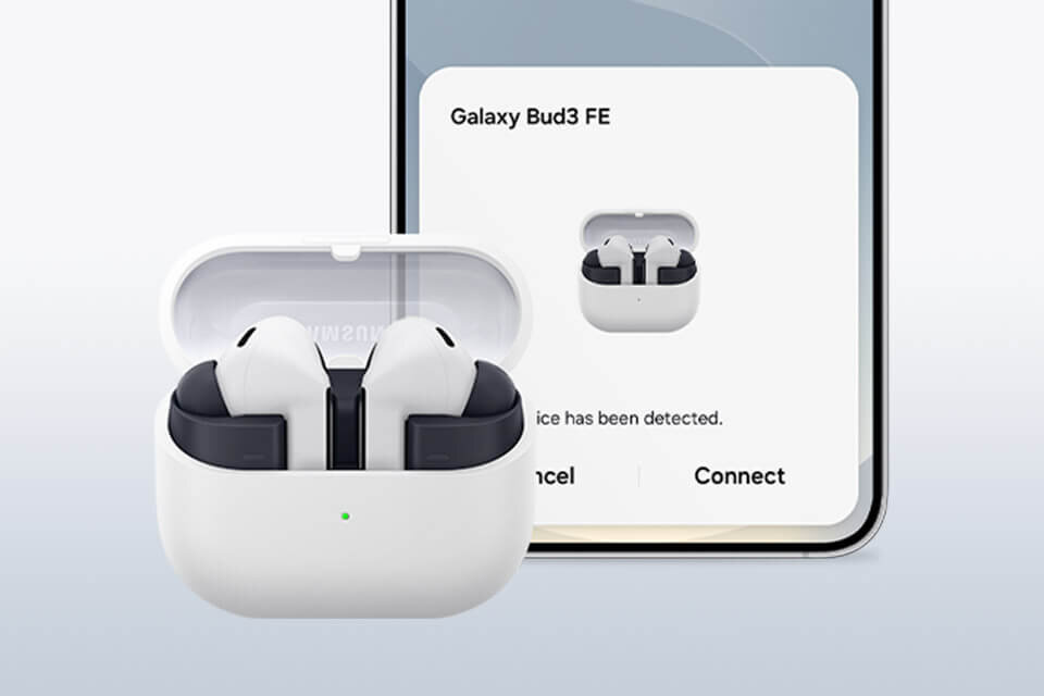Szare Galaxy Buds3 FE z otwartym etui ładującym, przed Samsungiem Galaxy S25, na którym wyświetlany jest ekran parowania. Na obrazie widoczne są słuchawki dokanałowe w białym etui oraz ekran telefonu z komunikatem \'Galaxy Bud3 FE\' i opcjami \'Cancel\' oraz \'Connect\'.