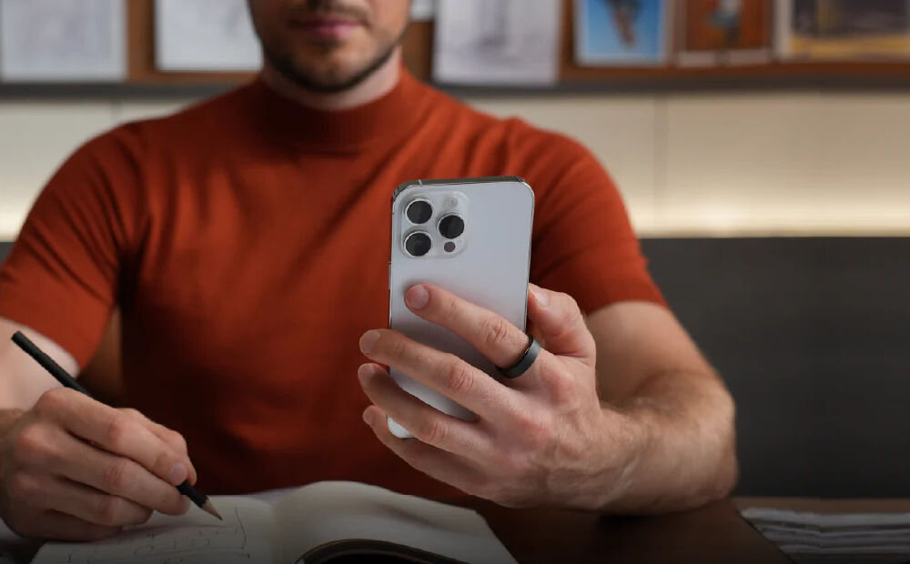 Osoba w czerwonej koszulce trzyma smartfon z czterema kamerami i nosi srebrny pierścień na palcu. Na stole leży otwarty notes, a w drugiej ręce znajduje się długopis.