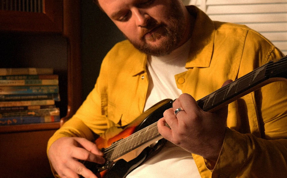 Osoba w żółtej koszuli gra na gitarze elektrycznej, nosząc srebrny pierścień na palcu. W tle widoczne są książki na półce.