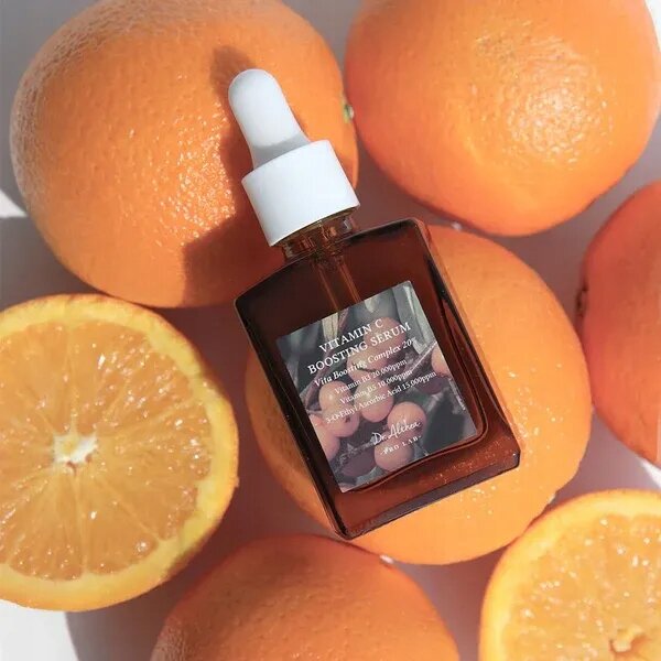 Na obrazie widoczna jest butelka z pipetą, na której etykiecie znajduje się napis \'Vitamin C Boosting Serum\'. W tle znajdują się całe i przekrojone pomarańcze.