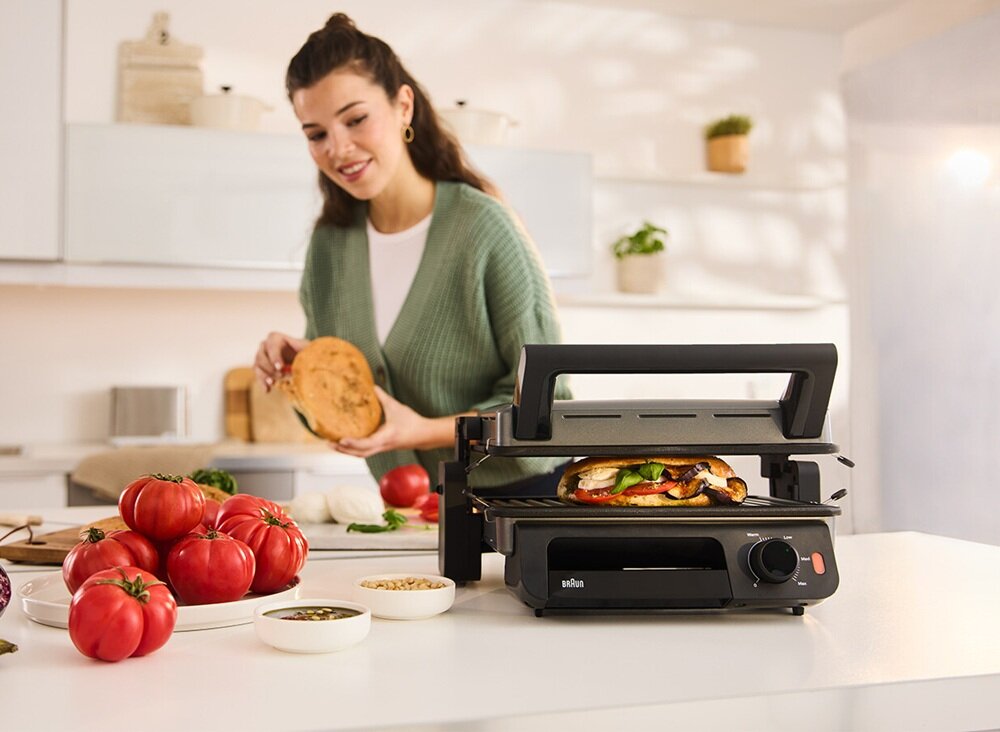 Na blacie kuchennym stoi czarny grill elektryczny z logo \'braun\', w którym znajduje się kanapka. Obok grilla leżą pomidory i miseczki z przyprawami, a w tle widać osobę przygotowującą jedzenie.