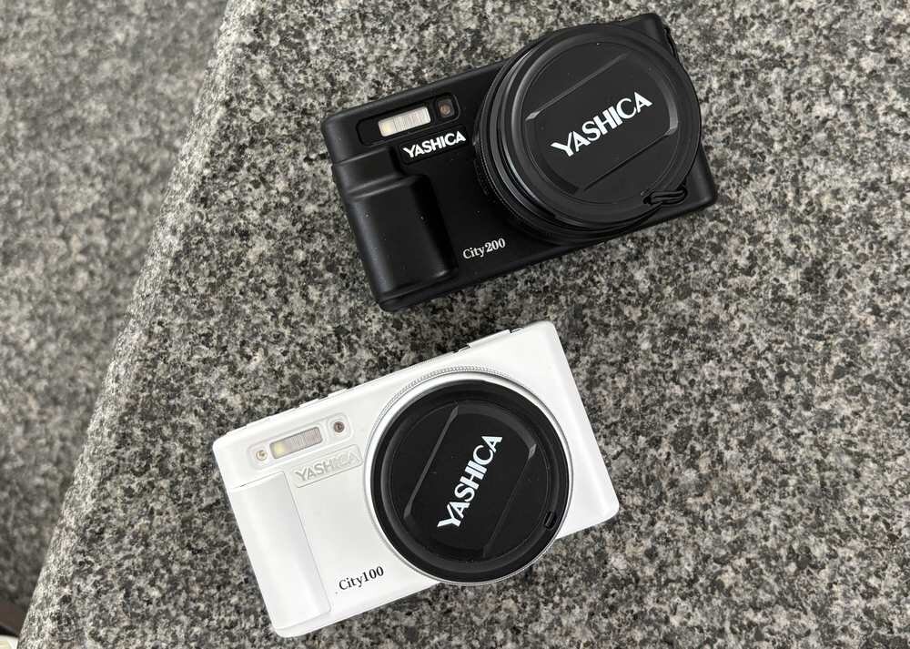 Na obrazie znajdują się dwa aparaty fotograficzne marki YASHICA, jeden czarny z napisem \'City 200\', a drugi biały z napisem \'City 100\'. Oba aparaty mają osłony obiektywów z logo \'YASHICA\'.