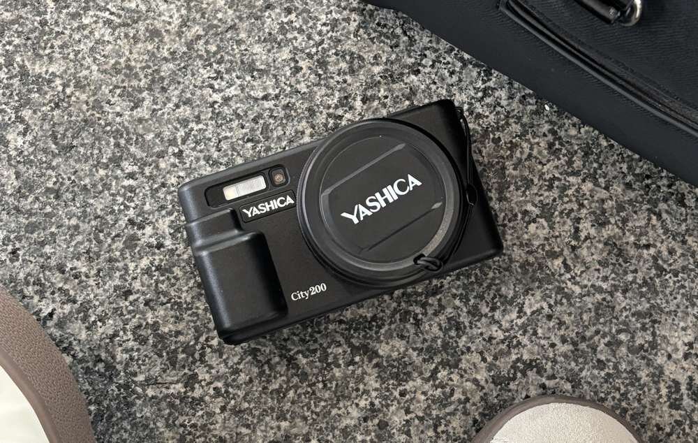 Czarny aparat fotograficzny z napisem \'YASHICA\' na osłonie obiektywu i korpusie, leży na powierzchni o teksturze przypominającej kamień. Obok aparatu widoczna jest część torby i fragment buta.