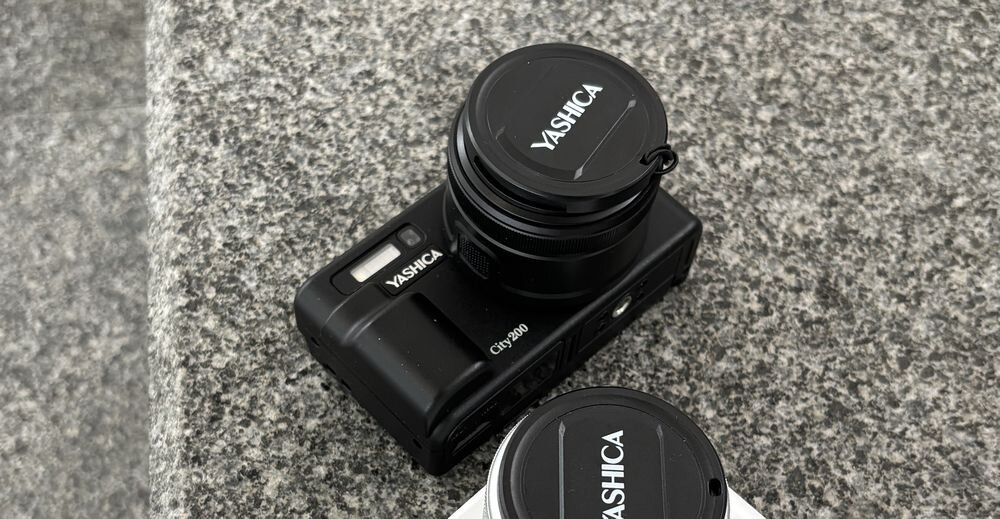 Czarny aparat fotograficzny z logo \'YASHICA\' na osłonie obiektywu, umieszczony na szarej powierzchni. Obok znajduje się drugi aparat z podobnym logo.