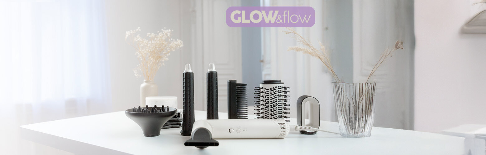 Na białym stole znajduje się zestaw akcesoriów do stylizacji włosów, w tym suszarko-lokówka i różne końcówki. W tle widoczny jest napis \'GLOW&flow\'.