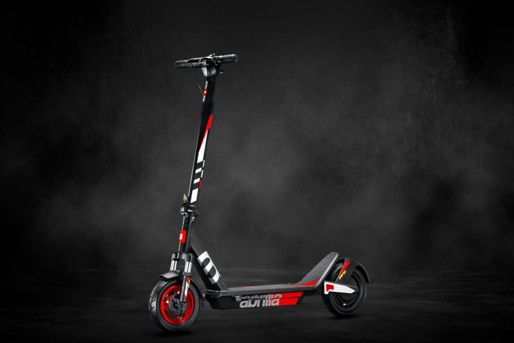 Czarno-szaro-czerwona hulajnoga elektryczna z widocznym logo \'aprilia\' na podeście, ustawiona na ciemnym tle. Hulajnoga ma czerwone akcenty na kołach i kierownicy.