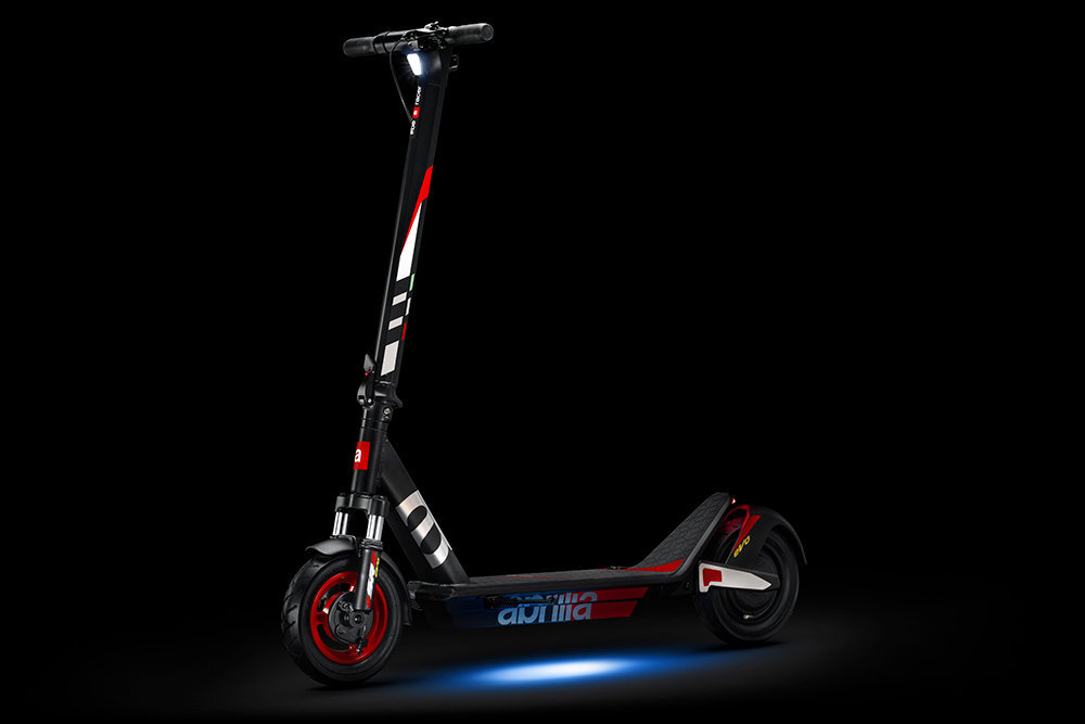 Czarno-szaro-czerwona hulajnoga elektryczna z widocznym logo \'aprilia\' na podeście. Posiada oświetlenie z przodu i niebieskie światło pod spodem.