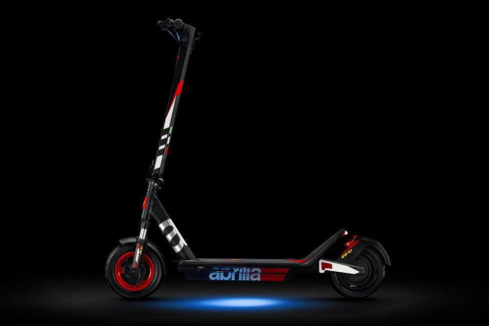 Czarno-szaro-czerwona hulajnoga elektryczna z widocznym logo \'aprilia\' na podeście. Hulajnoga ma czerwone akcenty na kołach i kierownicy, a tło jest ciemne.