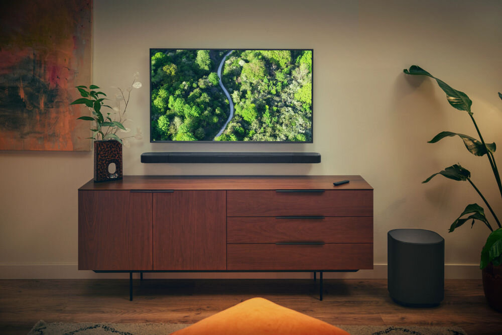 Na obrazie widać nowoczesny salon z telewizorem wyświetlającym zielony krajobraz, umieszczonym na ścianie nad drewnianą szafką. Pod telewizorem znajduje się soundbar, a obok szafki stoi czarny subwoofer.