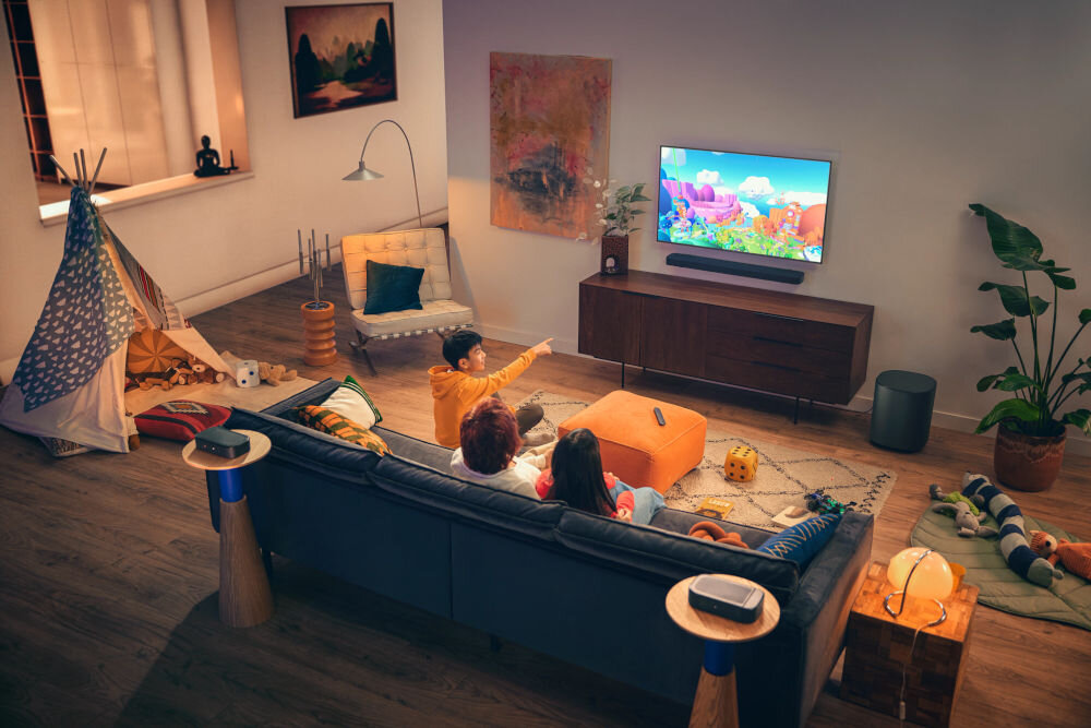 W salonie znajduje się telewizor na szafce, a przed nim siedzą trzy osoby na kanapie, oglądając kolorową animację. Obok telewizora umieszczony jest soundbar, a w pomieszczeniu widoczne są różne elementy dekoracyjne, takie jak rośliny i obrazy.