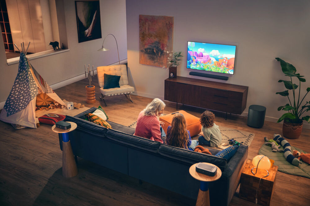 W salonie znajduje się telewizor na szafce, przed którym siedzą trzy osoby na kanapie. Pod telewizorem umieszczony jest soundbar, a obok szafki stoi bezprzewodowy subwoofer.