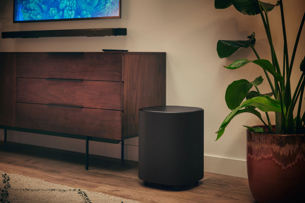 Na obrazie widać czarny subwoofer stojący obok drewnianej komody z trzema szufladami. Obok subwoofera znajduje się duża roślina doniczkowa.