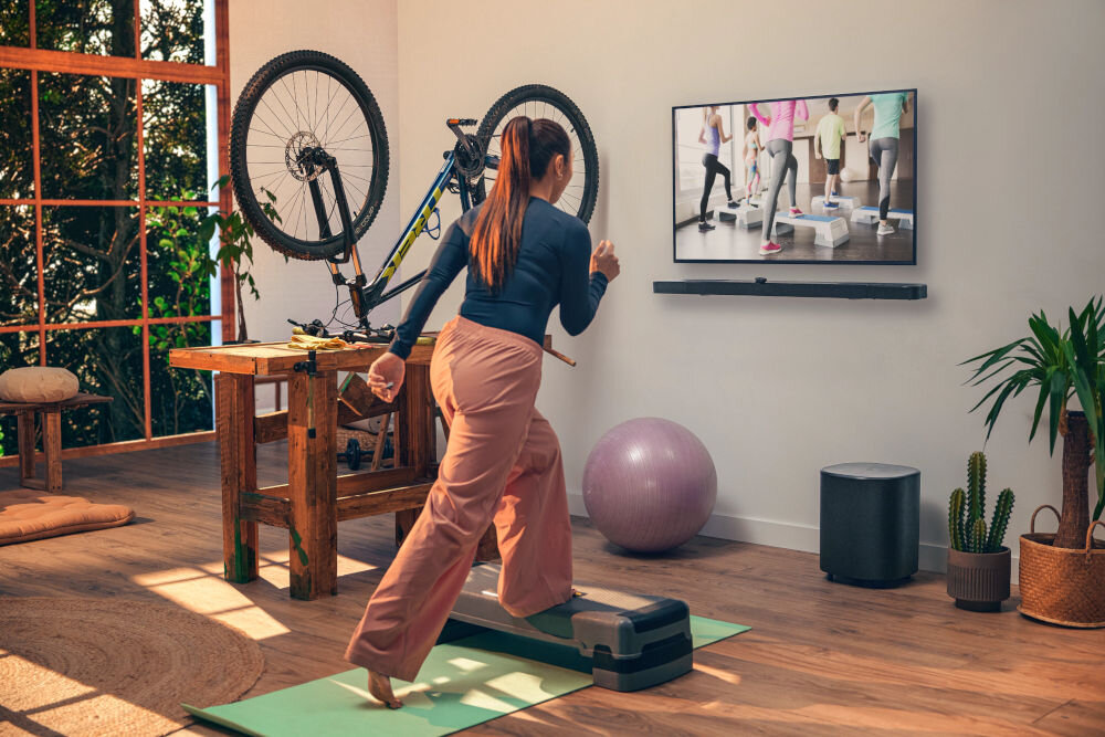 Osoba ćwiczy w salonie, patrząc na telewizor z programem fitness. Na ścianie pod telewizorem zamontowany jest soundbar, a obok stoi subwoofer.