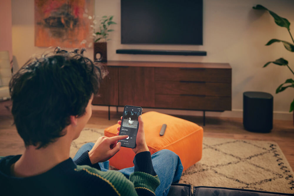 Osoba siedzi na kanapie, trzymając smartfon z aplikacją do sterowania dźwiękiem. W tle znajduje się telewizor z soundbarem i subwooferem na drewnianej szafce.