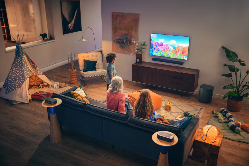 W salonie znajduje się telewizor na szafce, przed którym siedzą trzy osoby na kanapie. Obok telewizora umieszczony jest soundbar, a w pomieszczeniu widoczne są różne elementy dekoracyjne, takie jak rośliny i namiot dziecięcy.