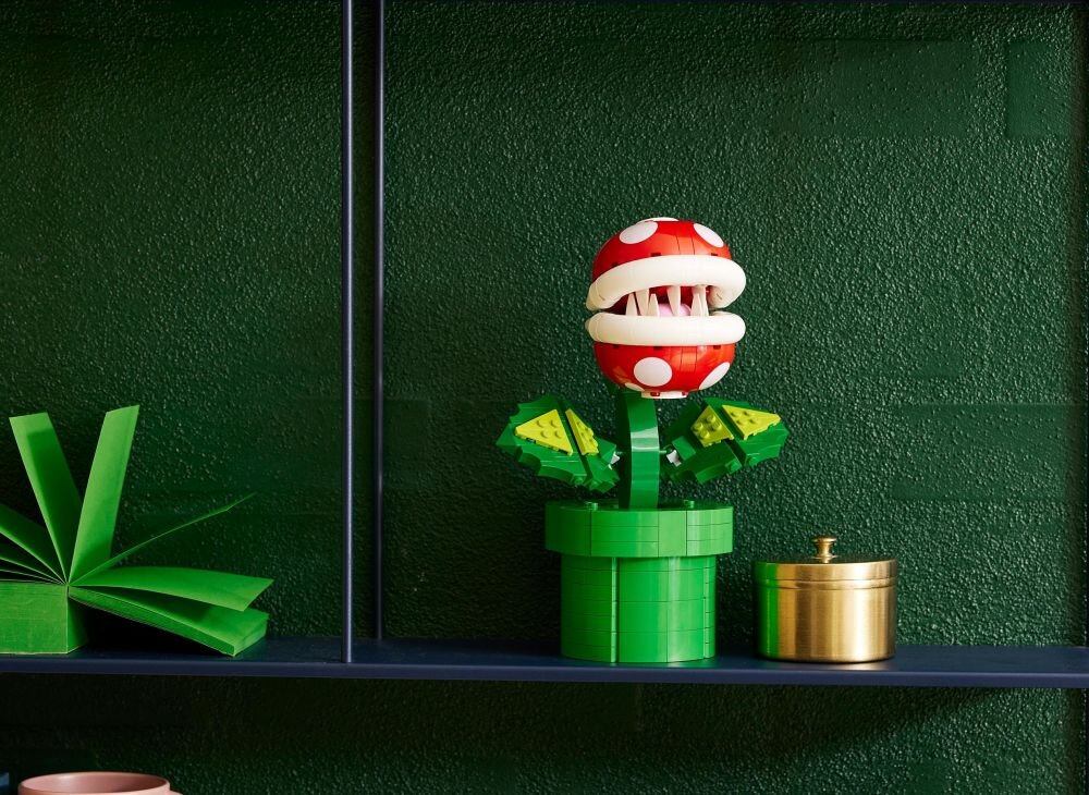 Na półce znajduje się model LEGO przedstawiający czerwony kwiatek z białymi kropkami i zębami, osadzony w zielonej doniczce. Obok stoi złoty pojemnik z pokrywką.