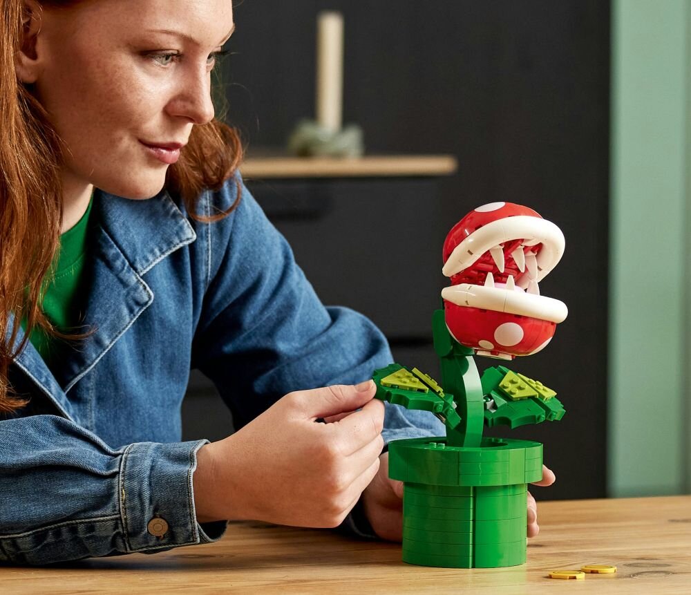 Osoba składa model LEGO przedstawiający roślinę z czerwonym kwiatem i białymi zębami, umieszczony w zielonej doniczce. Na stole leżą małe, żółte elementy LEGO.