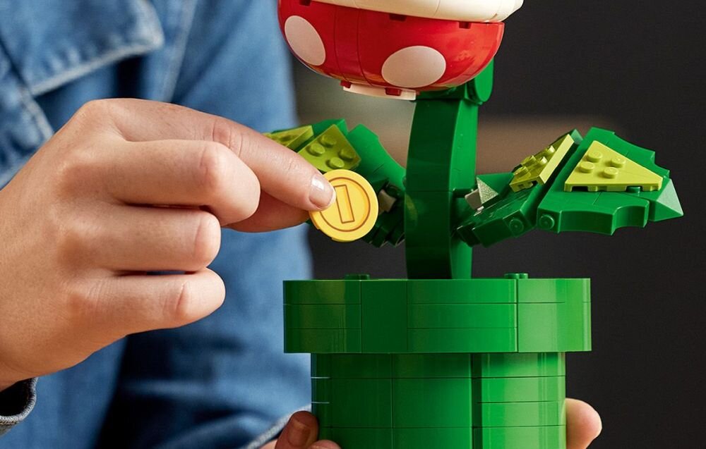 Osoba trzyma złotą monetę przy konstrukcji z klocków LEGO przedstawiającej roślinę z czerwonym kwiatem w doniczce. Roślina ma zielone liście i białe kropki na czerwonym kwiatku.