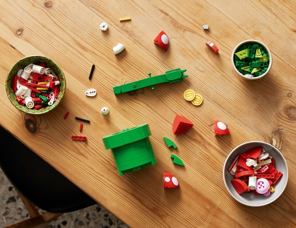 Na drewnianym stole leżą rozsypane klocki LEGO w różnych kolorach, w tym zielonym, czerwonym i białym. Obok znajdują się dwie miski z dodatkowymi elementami LEGO.