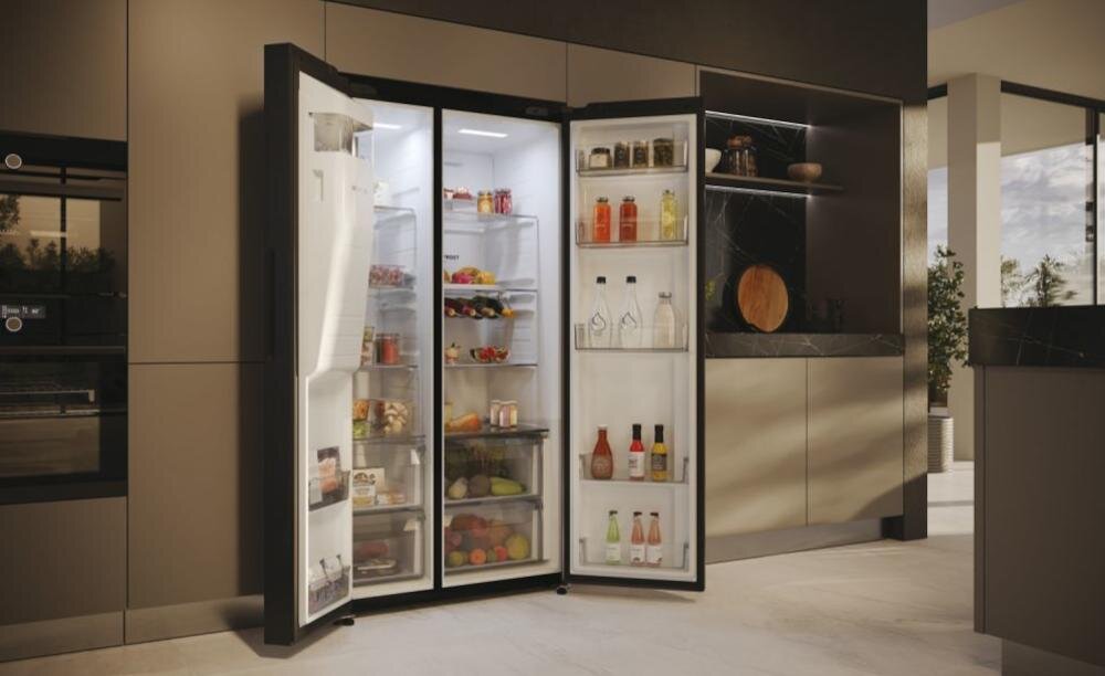 Otwarta lodówka w kuchni, wypełniona różnorodnymi produktami spożywczymi, takimi jak owoce, napoje i słoiki. Wnętrze lodówki jest dobrze oświetlone, a na drzwiach znajdują się dodatkowe półki z butelkami i kartonami.