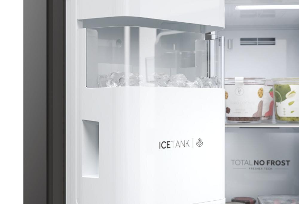 Na zdjęciu widoczna jest część lodówki z białym pojemnikiem na lód oznaczonym napisem \'ICE TANK\'. W tle widać otwartą lodówkę z produktami spożywczymi i napisem \'TOTAL NO FROST\'.