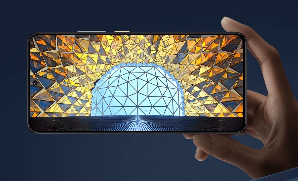 Osoba trzyma smartfon, na którego ekranie wyświetlany jest obraz z geometrycznym wzorem w odcieniach złota i niebieskiego. Tło jest jednolite i ciemne.