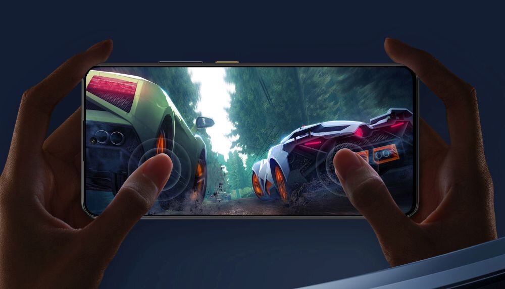Osoba trzyma smartfon, na ekranie którego widać wyścig dwóch samochodów sportowych na leśnej drodze. Samochody mają jaskrawe kolory i emitują efekty świetlne.