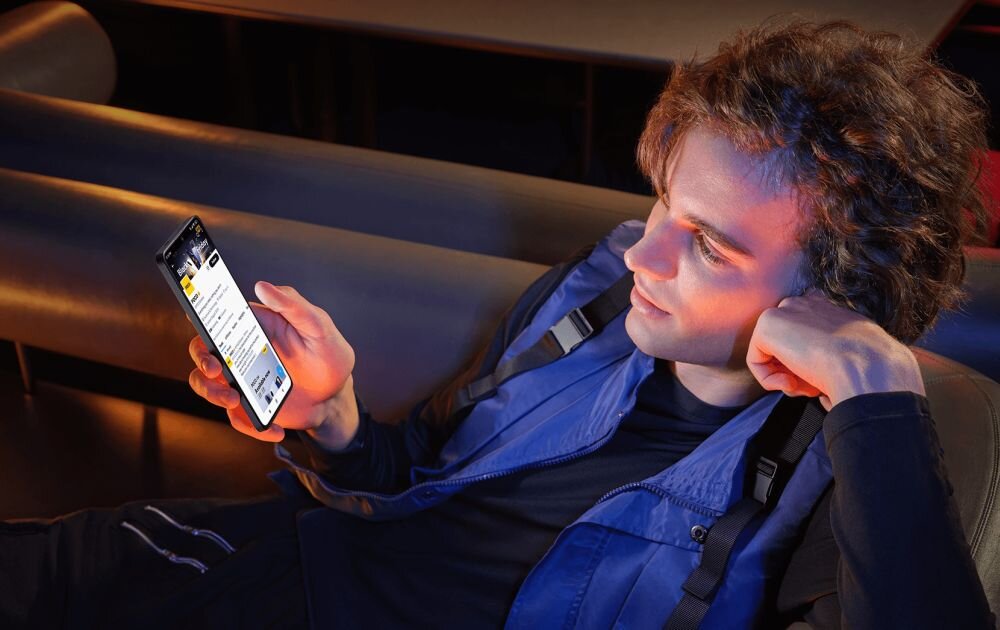 Osoba siedzi na kanapie, trzymając w ręku smartfon z widocznym ekranem. Na ekranie wyświetlana jest strona internetowa z tekstem i grafiką.