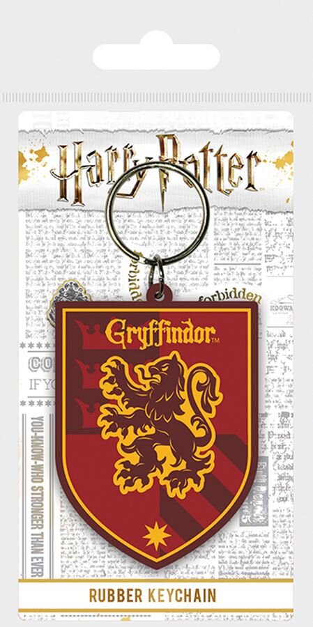 Gumowy brelok w kształcie herbu Gryffindoru z napisem \'Gryffindor\' i wizerunkiem lwa na czerwonym tle. W tle widoczny jest napis \'Harry Potter\' oraz fragmenty tekstu przypominające gazetę.