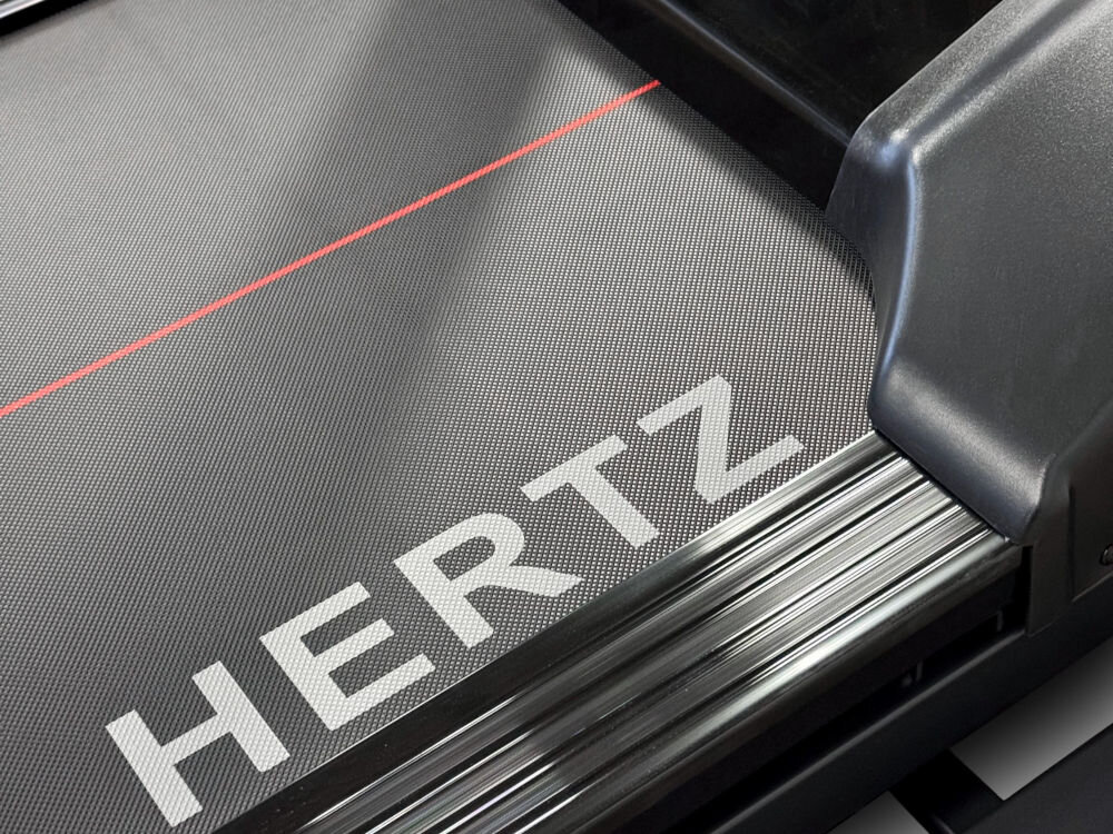 Fragment bieżni elektrycznej z widocznym napisem \'HERTZ\' na powierzchni bieżni oraz czerwoną linią. Elementy konstrukcyjne są w kolorze czarnym i metalicznym.