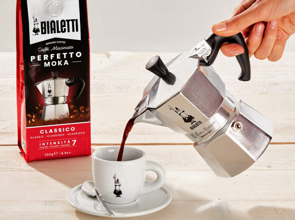 Osoba nalewa kawę z kawiarki do filiżanki, obok znajduje się opakowanie kawy z napisem \'BIALETTI Caffè Macinato PERFETTO MOKA CLASSICO INTENSITA 7\'.