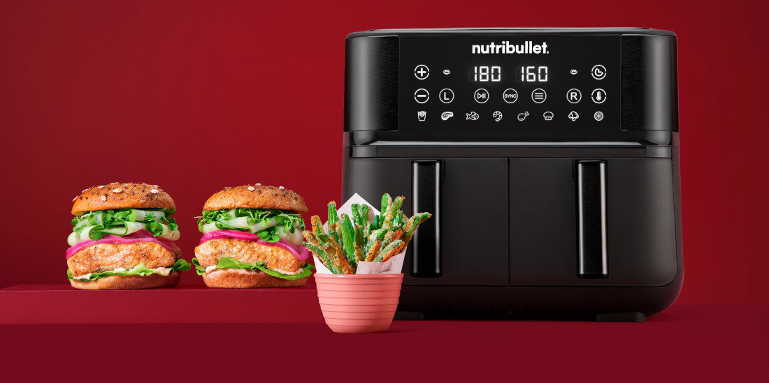 Na czerwonym tle znajduje się czarna frytkownica beztłuszczowa z wyświetlaczem i przyciskami dotykowymi, na którym widnieje logo \'nutribullet\'. Obok frytkownicy są dwa burgery i miseczka z frytkami warzywnymi.