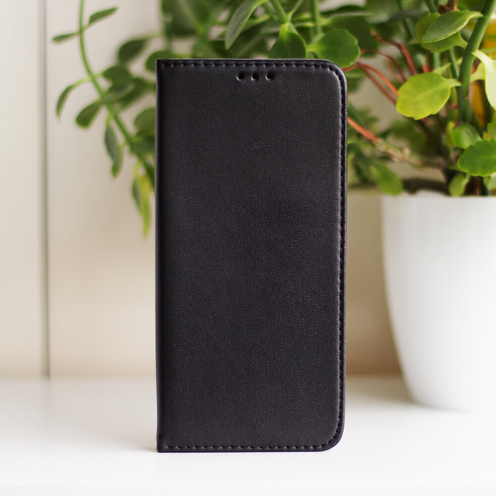Czarne etui na telefon w pozycji pionowej, wykonane z materiału przypominającego skórę, z trzema otworami na górze. W tle widoczne są zielone rośliny w białych doniczkach.